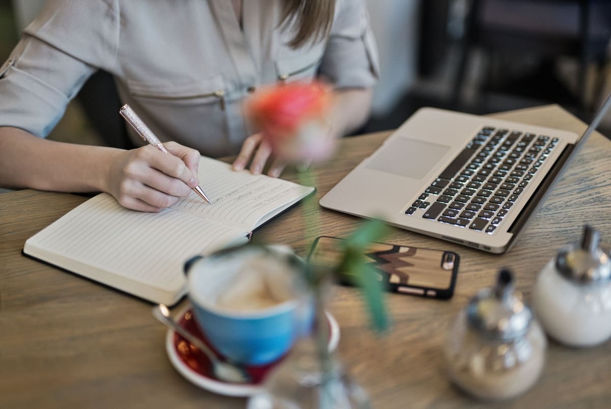 Blogger taking notes in a notebook beside a laptop and a cup of coffee