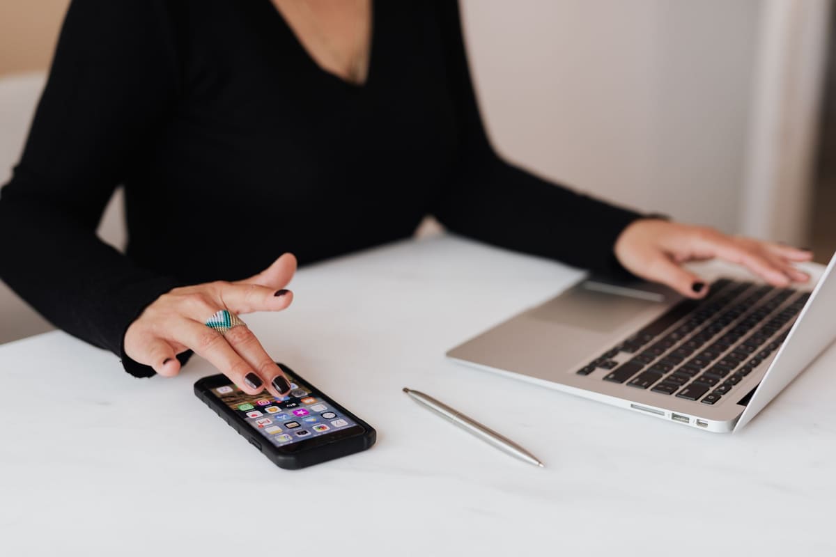 Person using a smartphone and a laptop at a white desk
