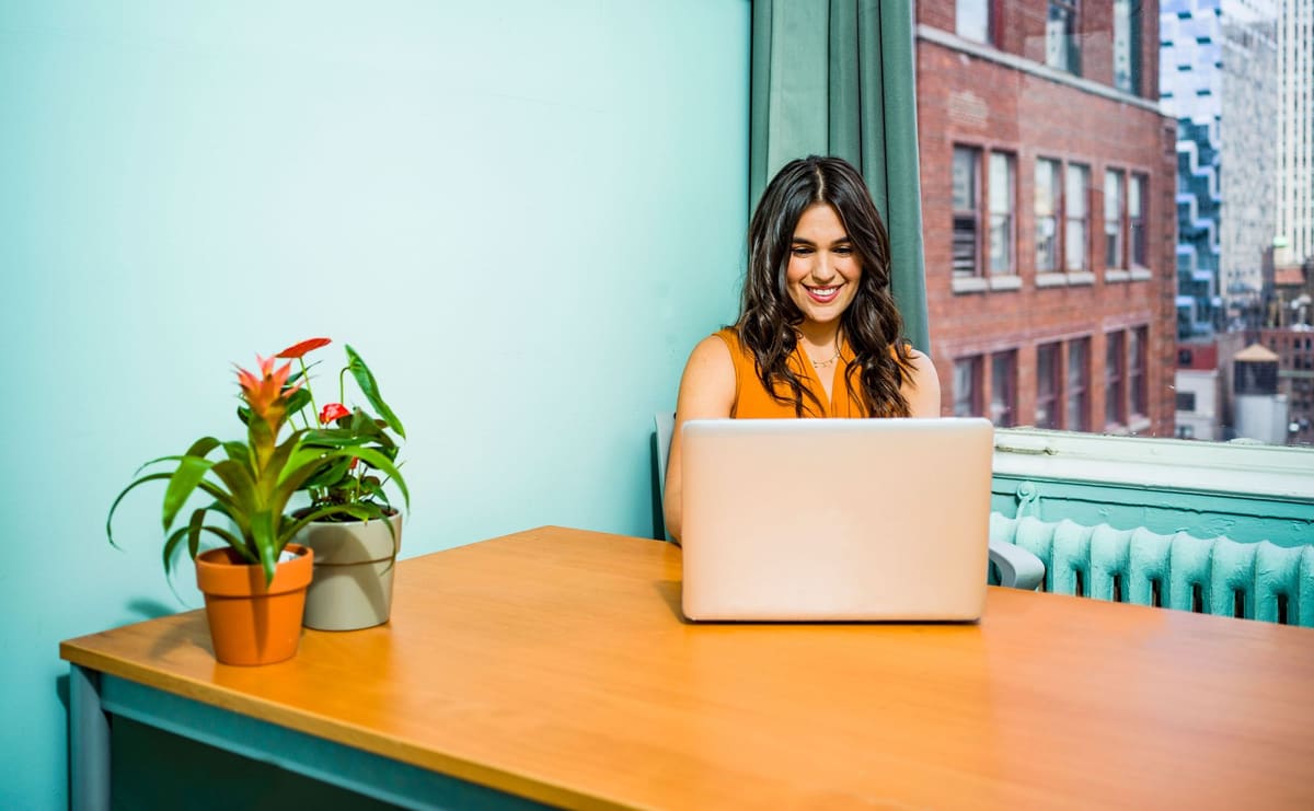 Smiling woman working on a laptop at a bright desk with plants by the window